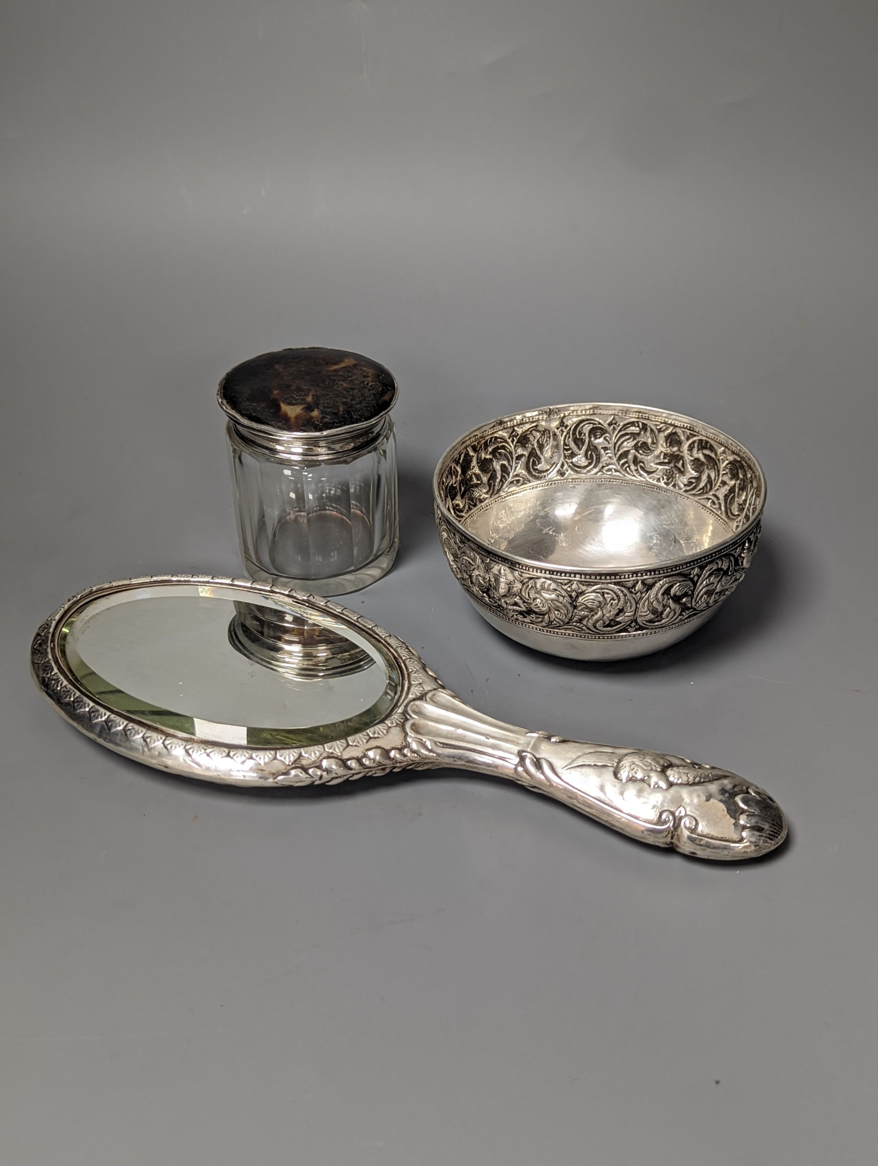 An Edwardian silver mounted 'Reynold's Angels' hand mirror, a silver and tortoiseshell lidded glass toilet jar and a Thai? white metal bowl.
